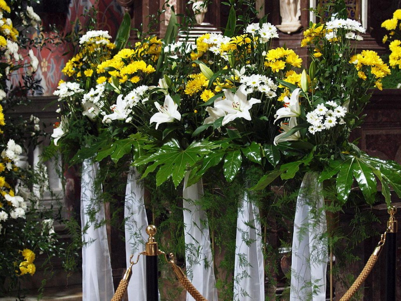 Dekoracja kwiatowa z białych lilii, żółtych i białych chryzantem, zielonych liści i białych wstążek, prawdopodobnie w kościele.