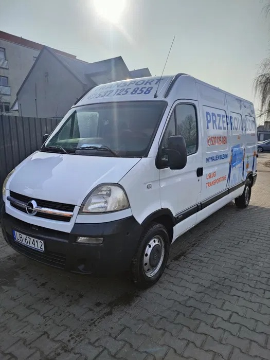 Biały bus z napisem 'Przeprowadzki' i numerem telefonu na tle brukowanej drogi i budynku. Widoczne logo marki Opel na masce.