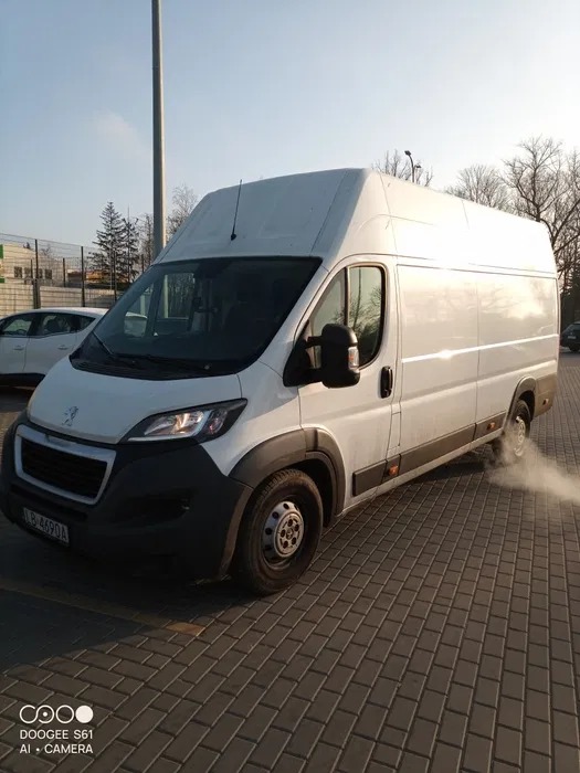 Biały bus Peugeot Boxer na kostce brukowej, z widocznym dymem z rury wydechowej, sugerujący gotowość do transportu. Słoneczny dzień, widok z przodu.