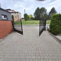Otwarta, dwuskrzydłowa brama wjazdowa z czarnego metalu na posesję z kostki brukowej, widok z zewnątrz. Brama z automatyką, zamontowana przy budynku.