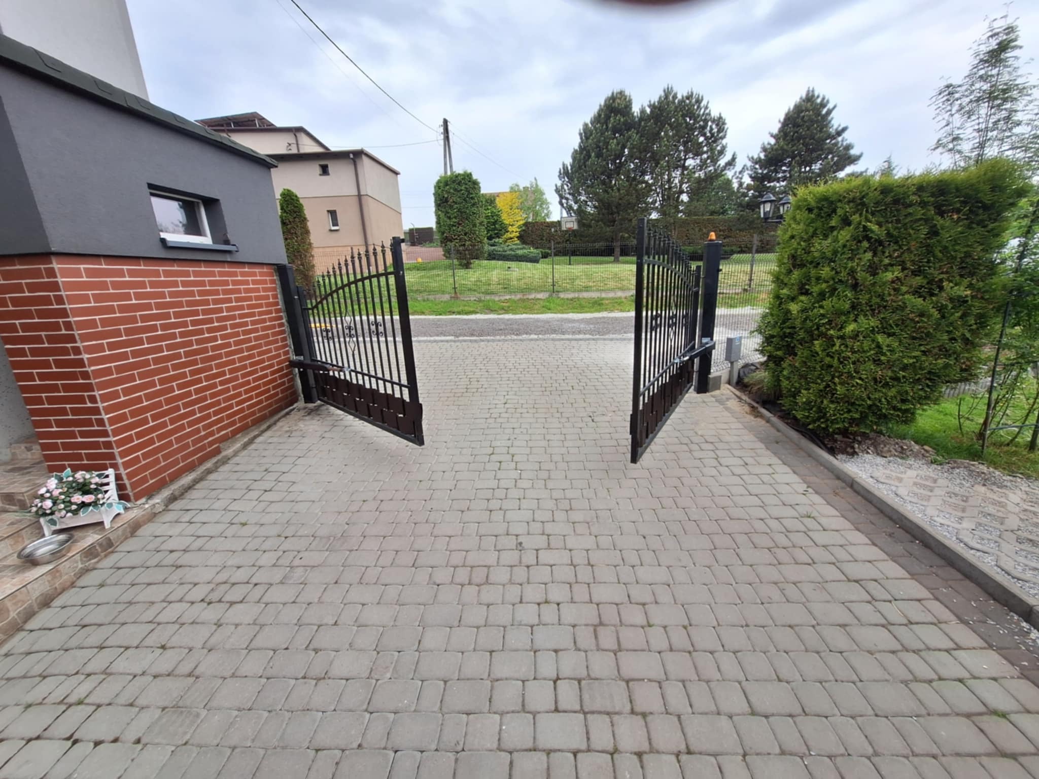 Otwarta, dwuskrzydłowa brama wjazdowa z czarnego metalu na posesję z kostki brukowej, widok z zewnątrz. Brama z automatyką, zamontowana przy budynku.