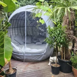Pokrowiec na kosz ogrodowy z przezroczystym frontem, wykonany na wymiar. W tle zieleń ogrodu i mały, biały pies. Zabezpieczenie przed wiatrem i wilgocią.