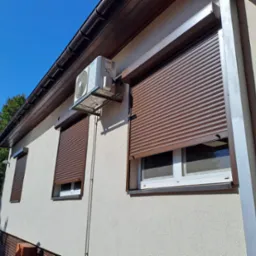 Elewacja domu z brązowymi roletami zewnętrznymi na oknach i klimatyzatorem. Widoczny fragment samochodu dostawczego.