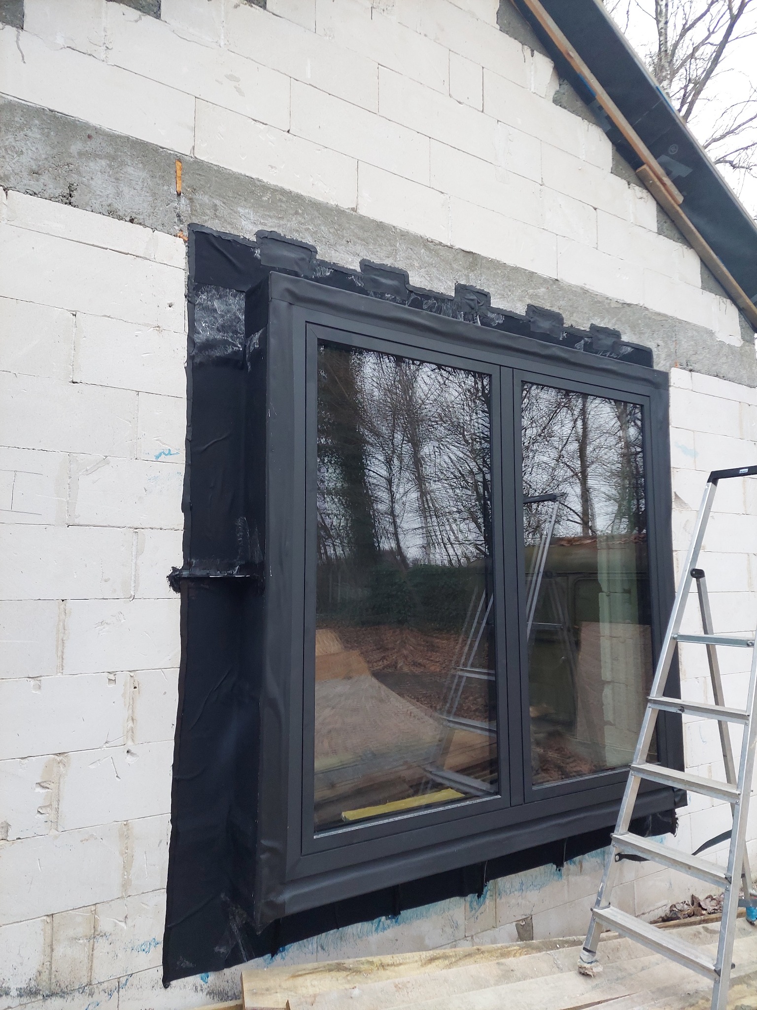 Montaż okna w surowym murze, z widoczną izolacją i drabiną. Okno dwuskrzydłowe z ciemną ramą, odbijające widok drzew. Etap prac wykończeniowych.