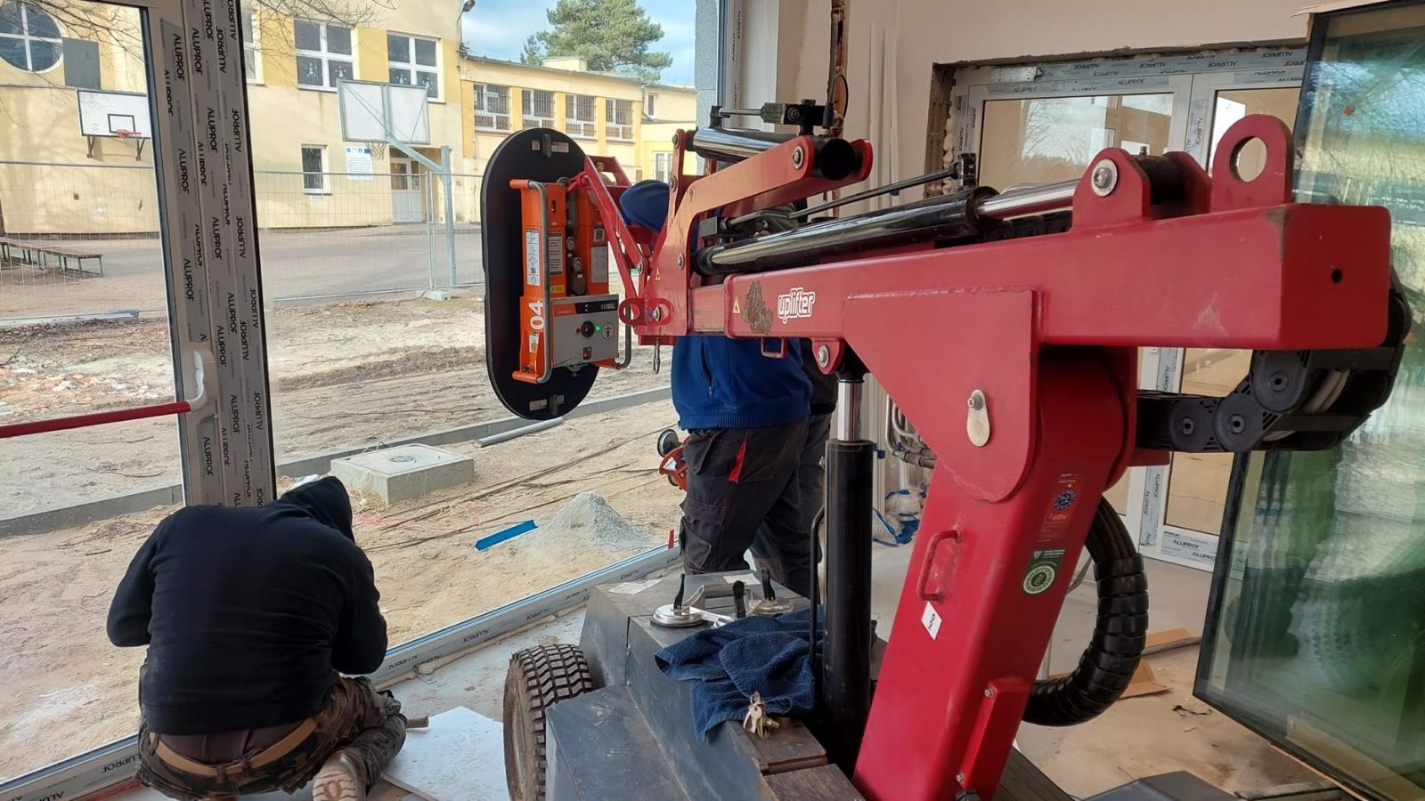 Montaż okna z użyciem specjalistycznego podnośnika UPLIFTER, pracownik w trakcie uszczelniania ramy, widoczne profile okienne ALLUPROF.