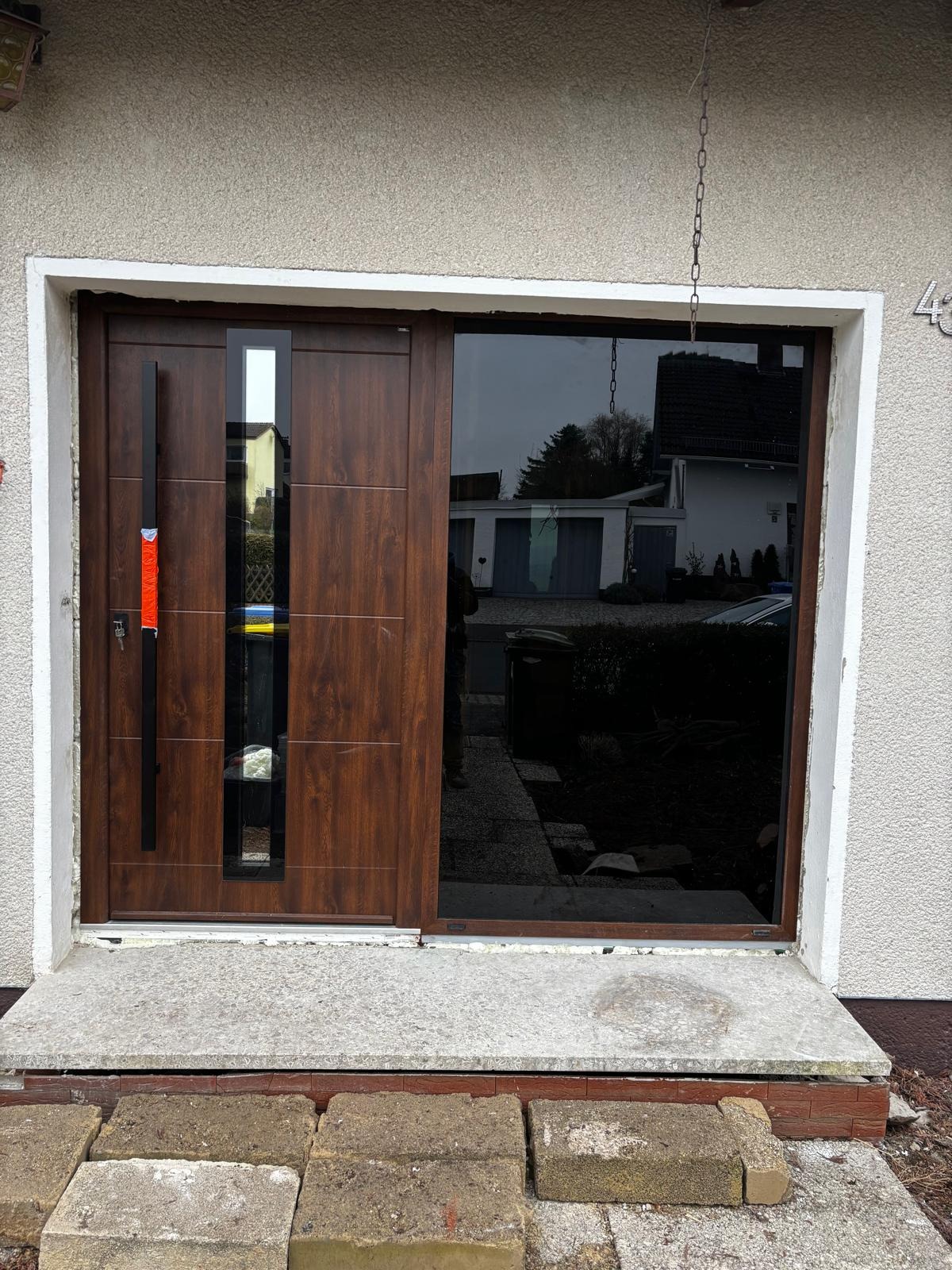 Brązowe drzwi wejściowe z szybą, czarny, długi uchwyt, częściowo widoczne cegły na progu. Dom z szarą elewacją, numer 43.