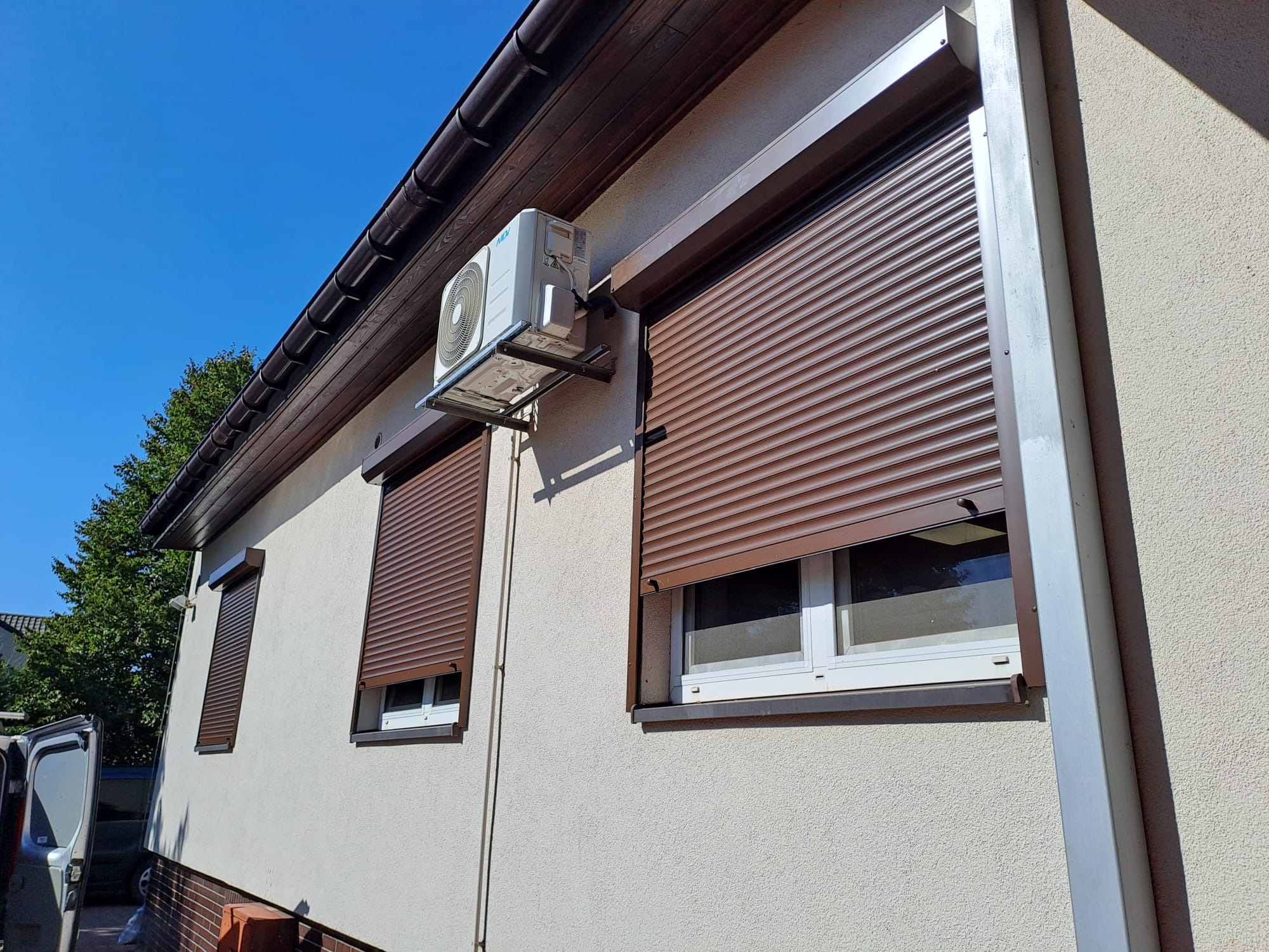 Elewacja domu z brązowymi roletami zewnętrznymi na oknach i klimatyzatorem. Widoczny fragment samochodu dostawczego.