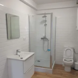 Jasna łazienka z kabiną prysznicową, umywalką z szafką i toaletą. Białe płytki na ścianach i drewnopodobna podłoga. Minimalistyczny design, nowoczesne wykończenie.