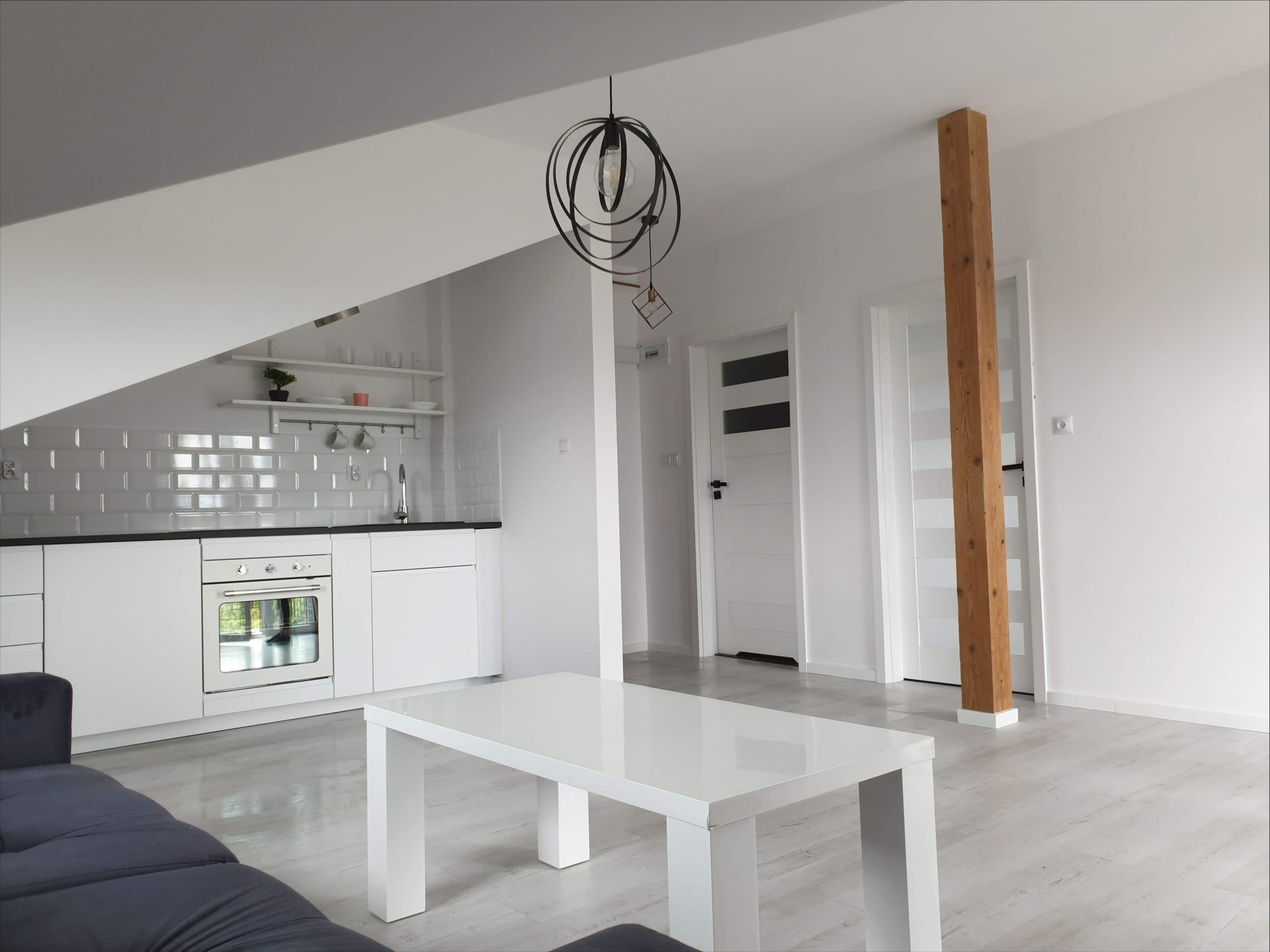 Jasne wnętrze po remoncie: biała kuchnia z szarą glazurą, drewniany filar i nowoczesna lampa nad białym stolikiem kawowym. Minimalistyczny design.