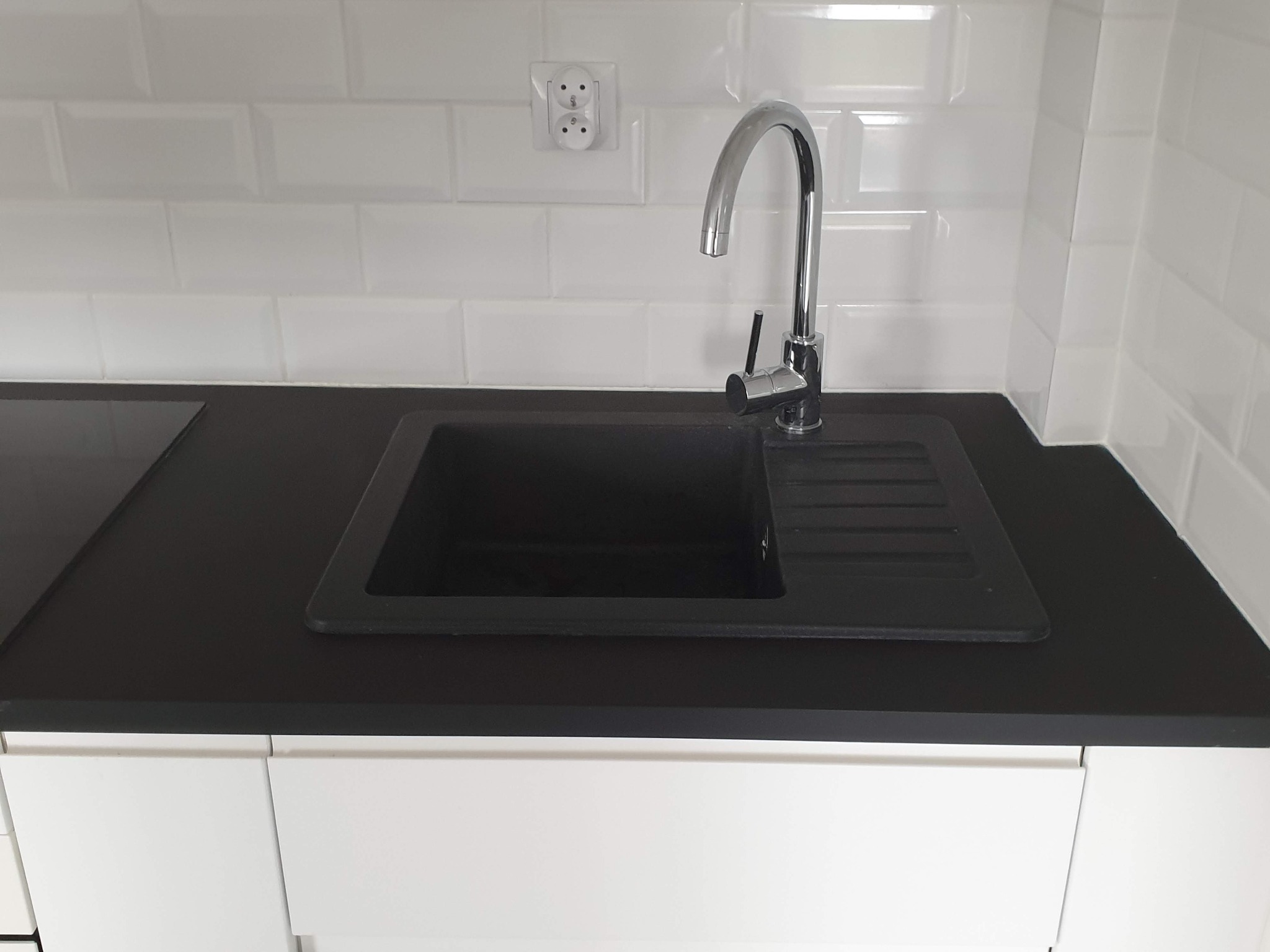 Nowoczesna, czarna umywalka z chromowaną baterią na tle białych, błyszczących płytek typu metro i białych szafek. Widoczne gniazdko elektryczne.