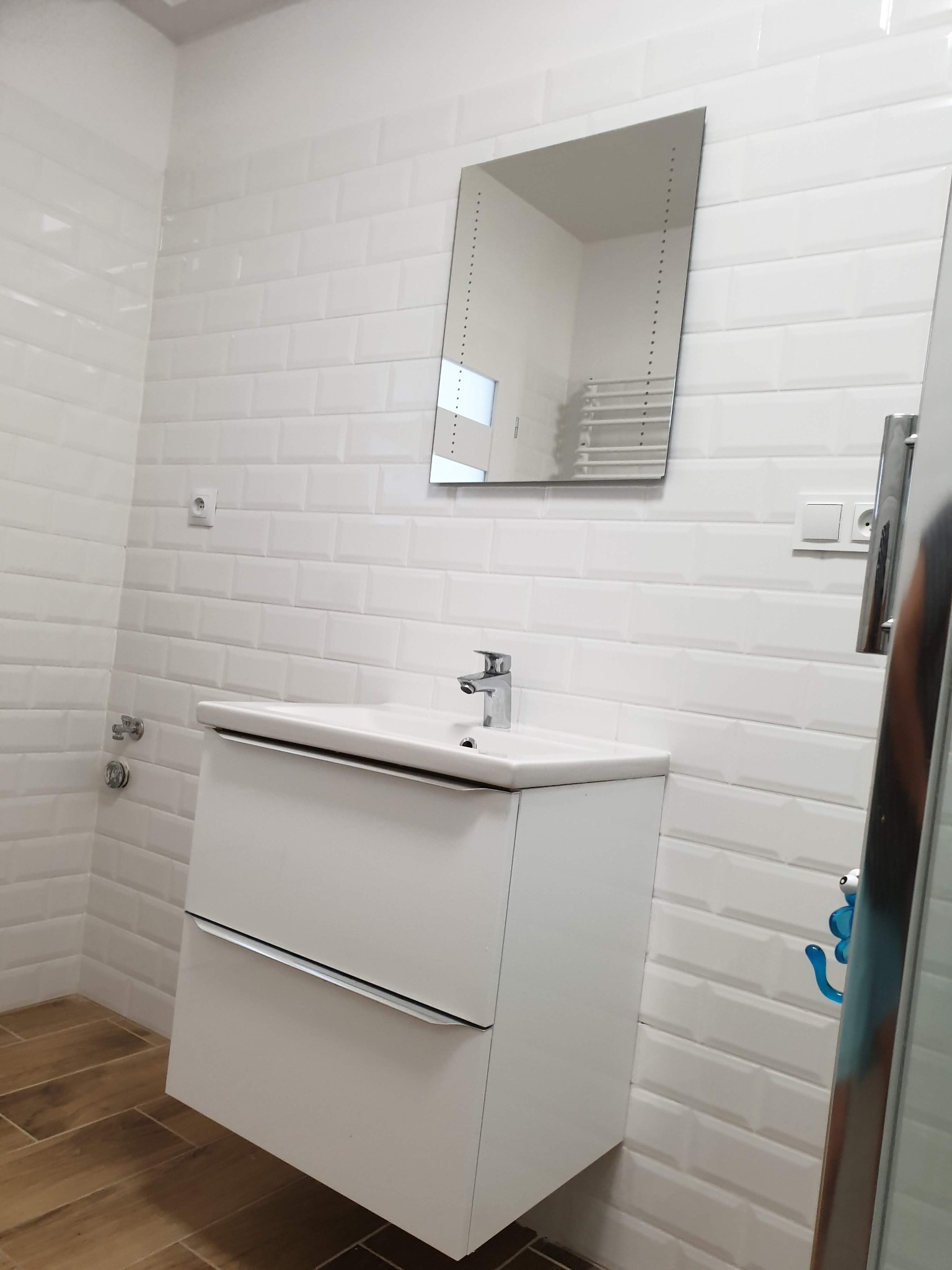 Biała umywalka z szafką w łazience z białymi, prostokątnymi płytkami na ścianach i lustrem z podświetleniem. Widoczna armatura i drewnopodobna podłoga.