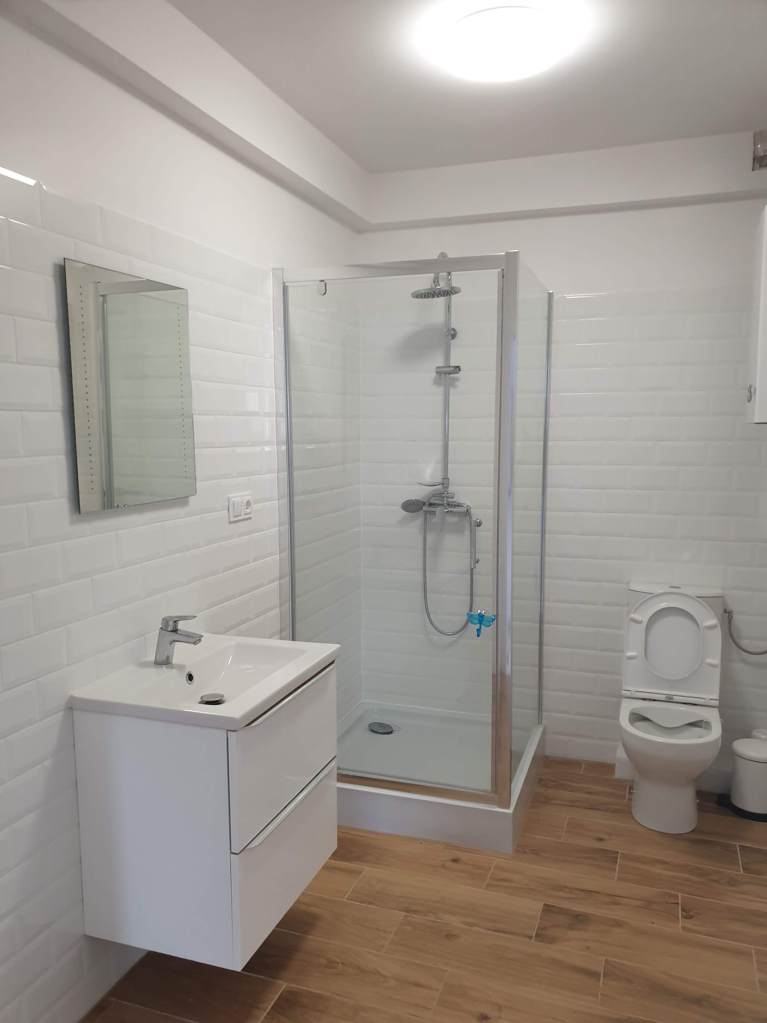 Jasna łazienka z kabiną prysznicową, umywalką z szafką i toaletą. Białe płytki na ścianach i drewnopodobna podłoga. Minimalistyczny design, nowoczesne wykończenie.