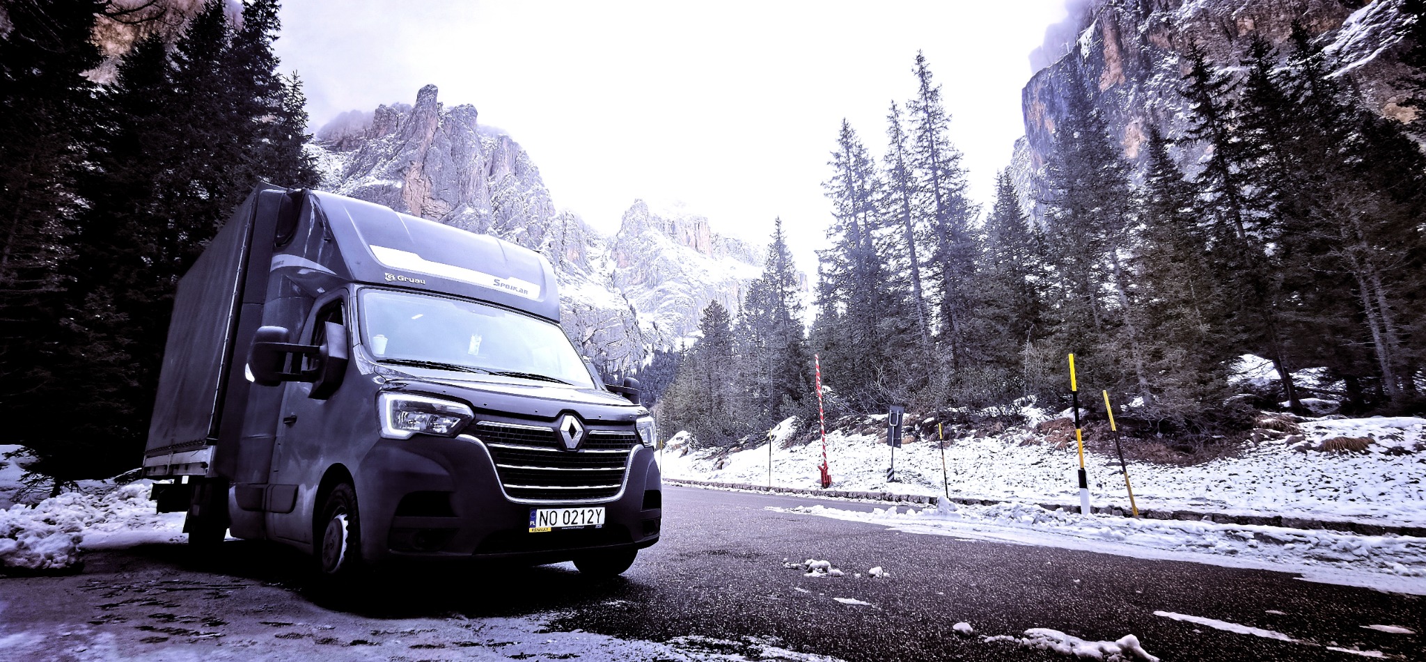 Czarny bus dostawczy Renault na tle górskiego krajobrazu w zimowej scenerii. Śnieg pokrywa pobocze drogi i drzewa. Widoczne tablice rejestracyjne.