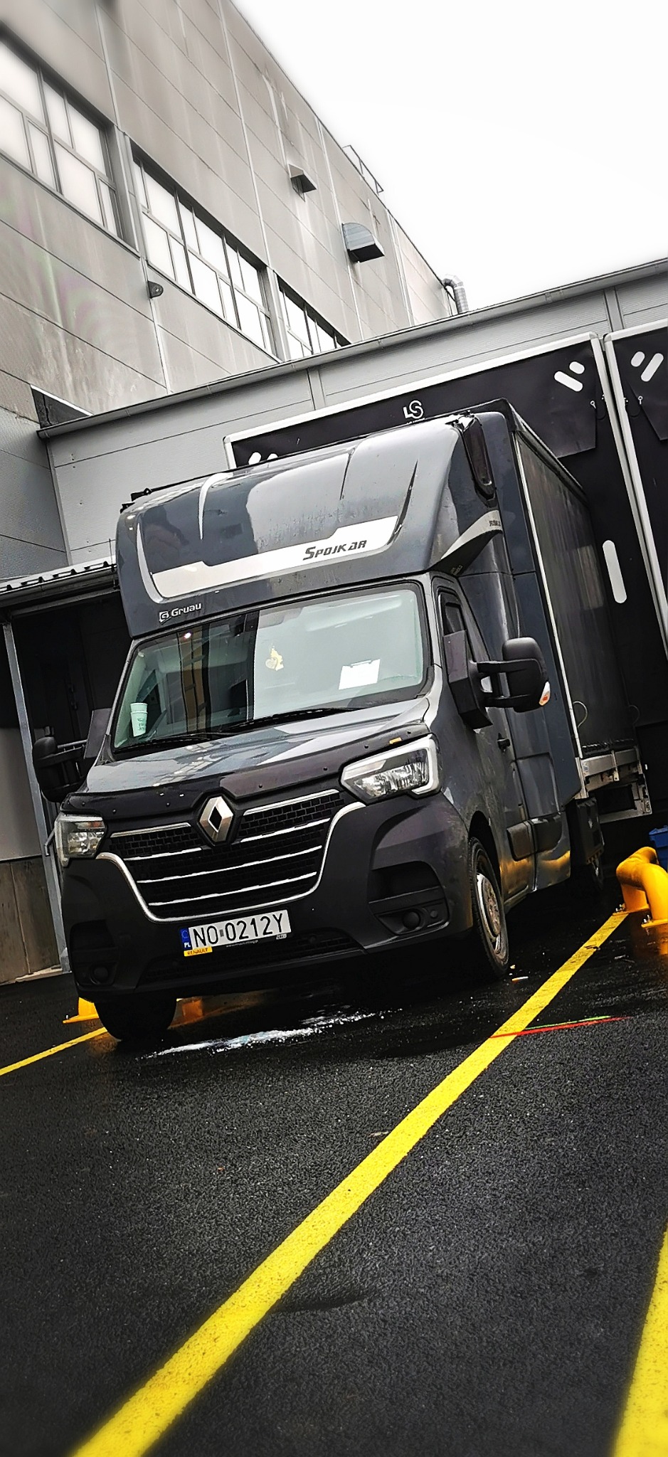 Szary bus Renault Master z zabudową Spojkar, zaparkowany na parkingu z żółtymi liniami, prawdopodobnie gotowy do załadunku lub rozładunku towaru.