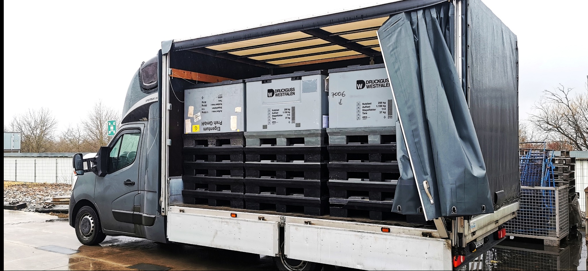 Ciężarówka z otwartą plandeką, załadowana skrzyniami na paletach. Widoczne oznaczenia firmowe na skrzyniach i detale konstrukcyjne pojazdu.