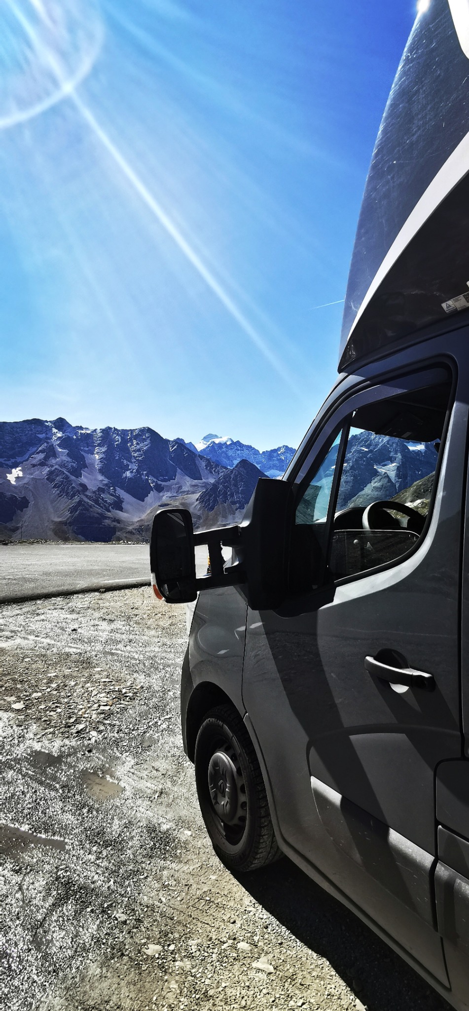 Szary bus z nadbudówką na tle górskiego krajobrazu w słoneczny dzień. Widoczny fragment pojazdu, w tle ośnieżone szczyty i błękitne niebo.