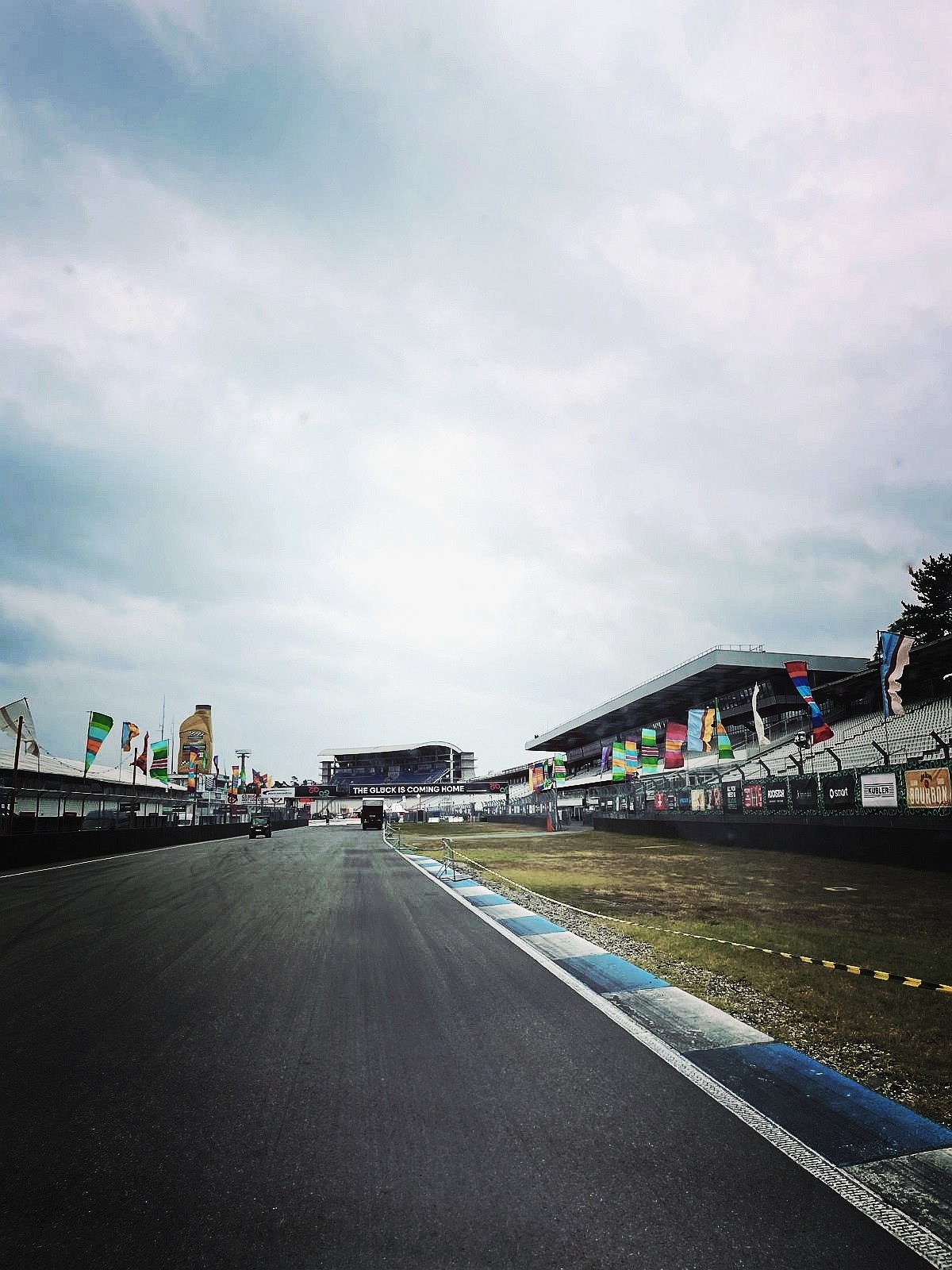 Pusty tor wyścigowy Hockenheimring z flagami, trybunami i wiaduktem z napisem 'The Glock is coming home' w pochmurny dzień. Widok z perspektywy kierowcy.