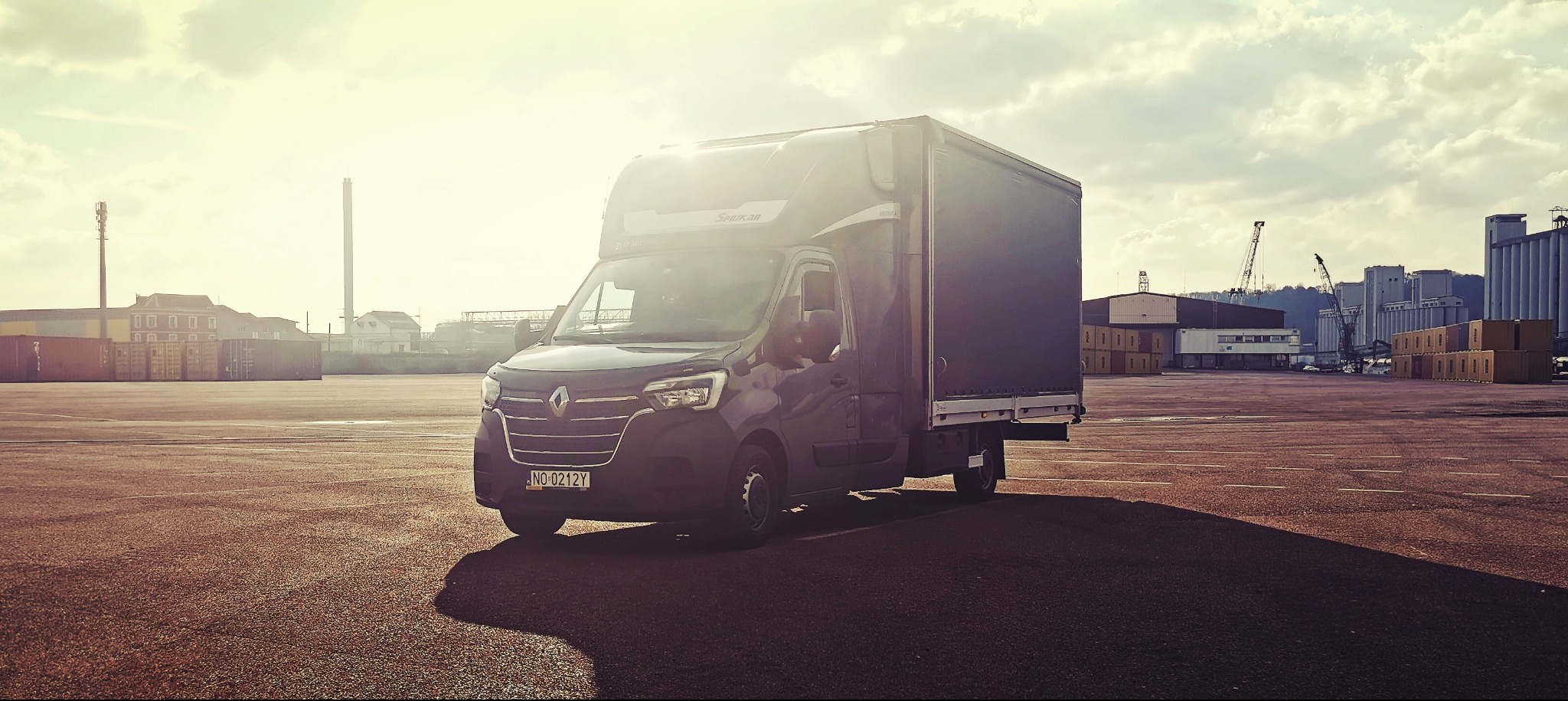 Czarny bus dostawczy Renault Master z zabudową skrzyniową na placu portowym, w tle kontenery, żurawie i budynki przemysłowe, słoneczny dzień.
