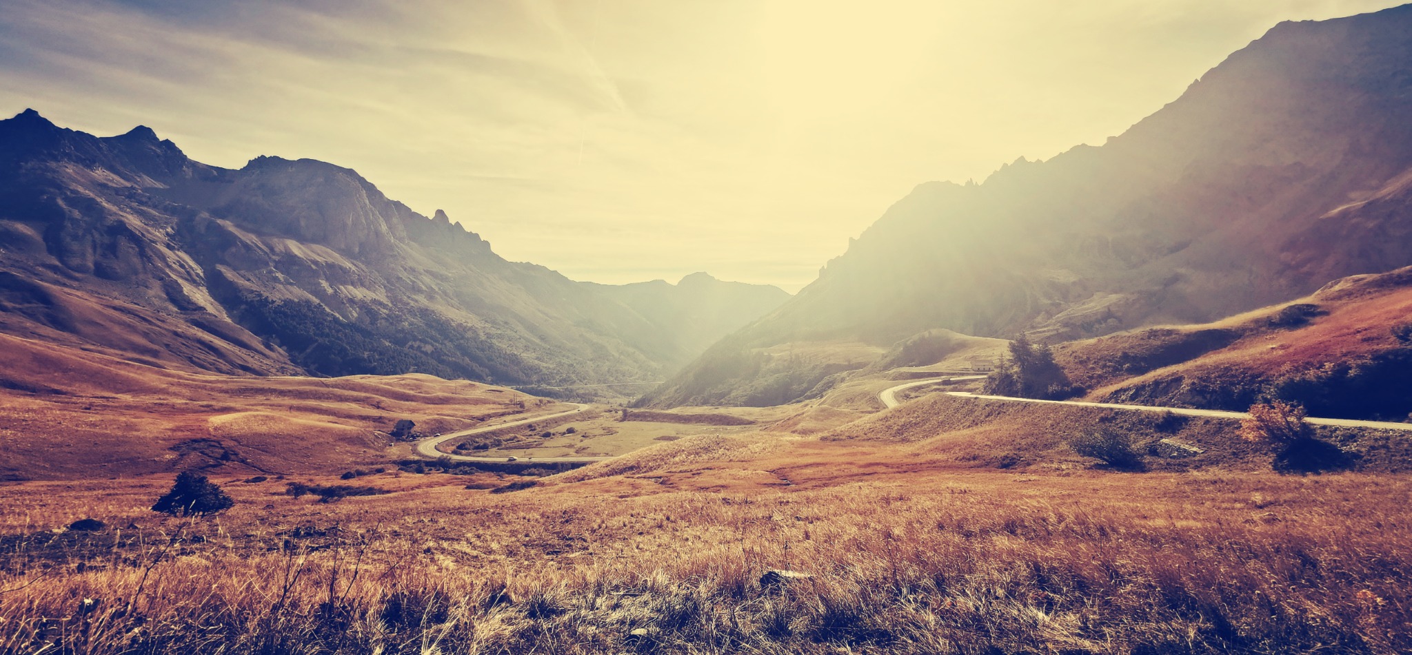 Krajobraz górski z krętą drogą w dolinie, oświetlony ciepłym światłem słonecznym. Widok z perspektywy trawy, charakterystyczne kolory jesieni.