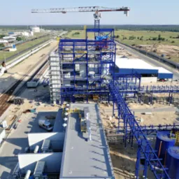 Widok z góry na budowę zakładu przemysłowego z konstrukcją stalową w kolorze niebieskim, dźwigiem i elementami infrastruktury kolejowej w tle.