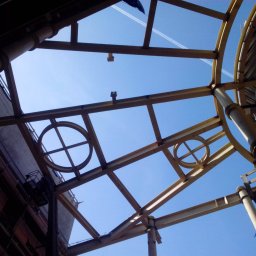 Ziemowit Otto - Konstrukcja stalowa dachu z okrągłymi elementami dekoracyjnymi, widziana z dołu na tle błękitnego nieba z smugami kondensacyjnymi samolotów.