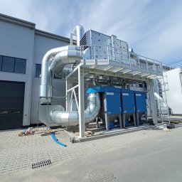 Ziemowit Otto - Zewnętrzna instalacja przemysłowa z srebrnymi rurami i niebieskimi filtrami, zamontowana przy szarej hali z bramą garażową, widoczna w słoneczny dzień.