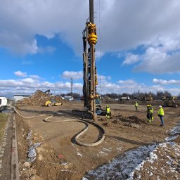 Ziemowit Otto - Prace ziemne w toku: ekipa w żółtych kamizelkach nadzoruje wbijanie pali w grunt za pomocą palownicy. W tle widoczny sprzęt budowlany i błękitne niebo z chmurami.