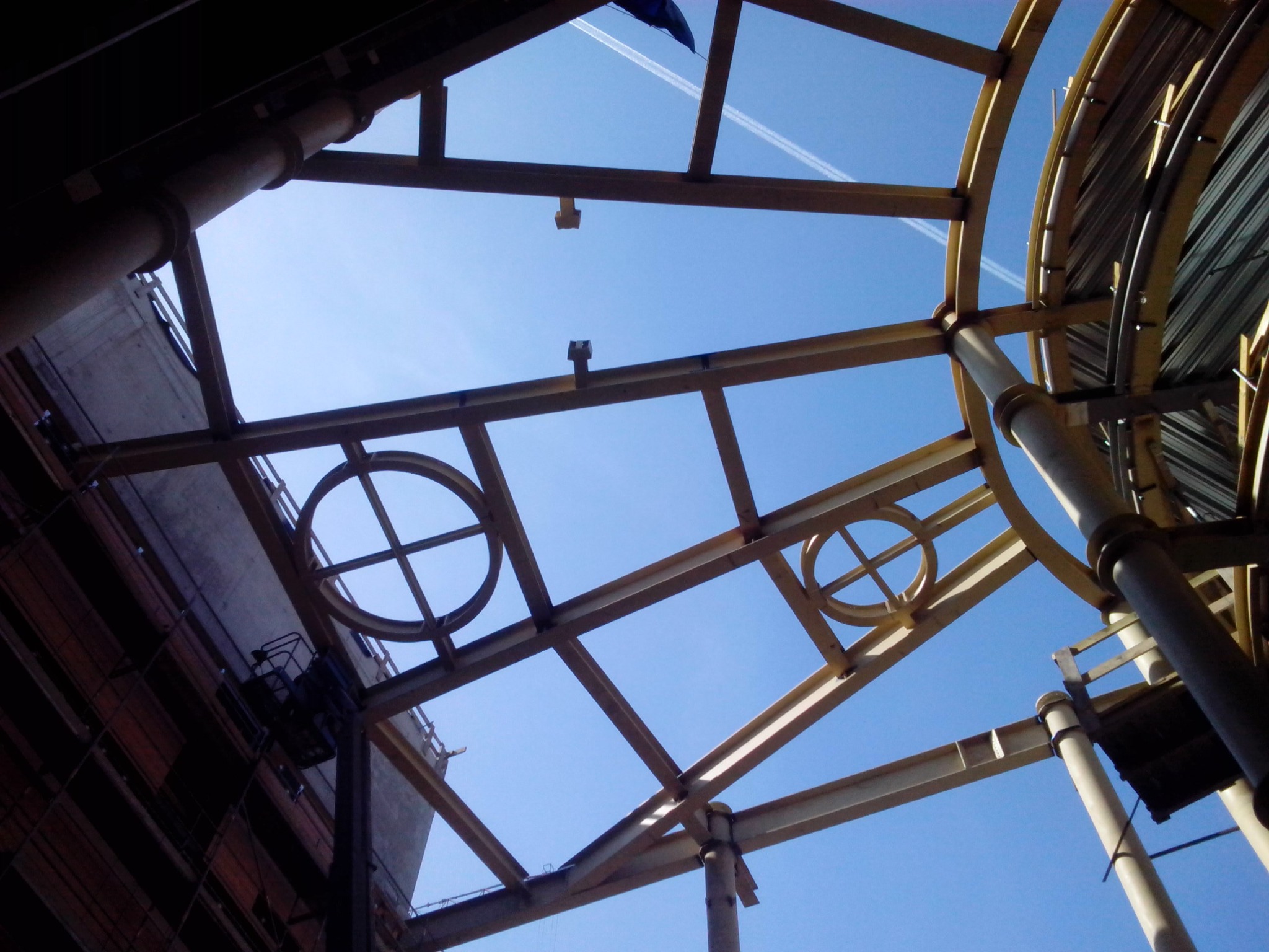 Konstrukcja stalowa dachu z okrągłymi elementami dekoracyjnymi, widziana z dołu na tle błękitnego nieba z smugami kondensacyjnymi samolotów.