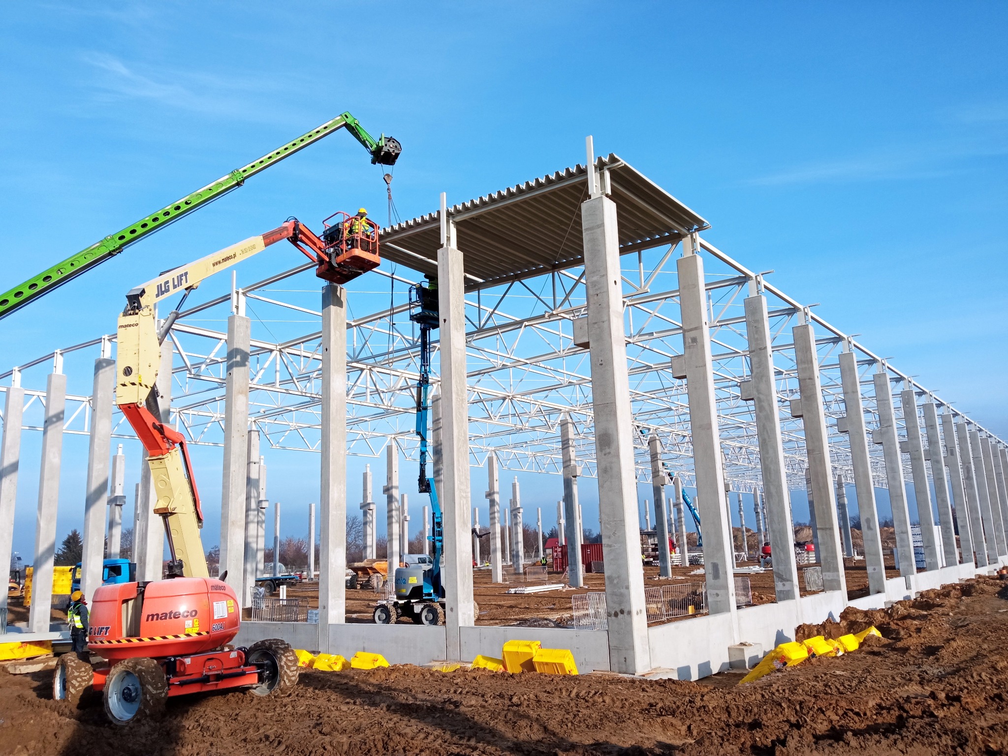 Montaż konstrukcji stalowej hali magazynowej z użyciem podnośników koszowych na tle błękitnego nieba. Widoczne betonowe słupy i stalowe elementy dachu.
