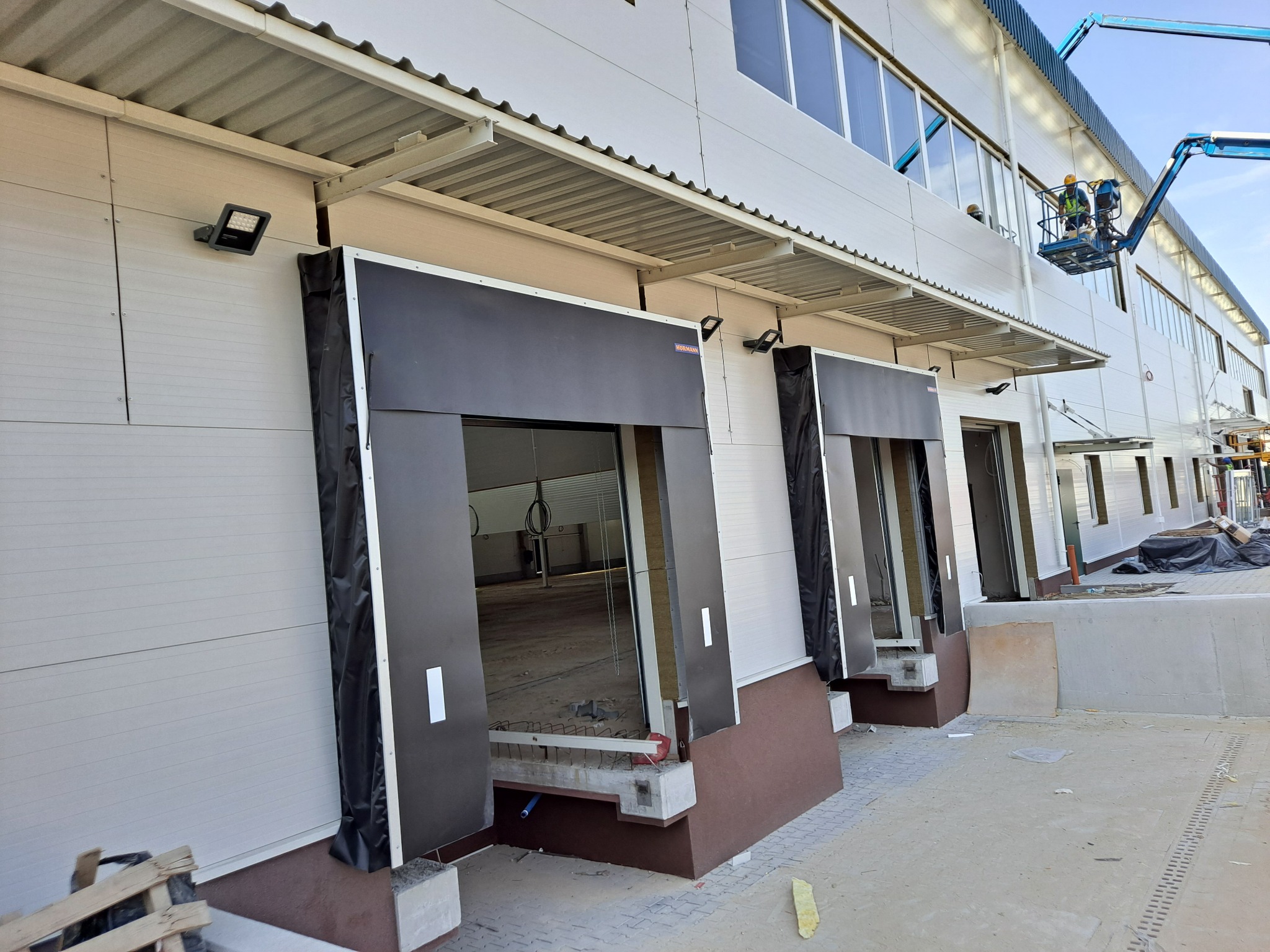 Montaż bram przemysłowych w nowym budynku magazynowym. Widoczny podnośnik z pracownikami na tle jasnej elewacji i otwartych wrót. Detal: zabezpieczenia na bramach.