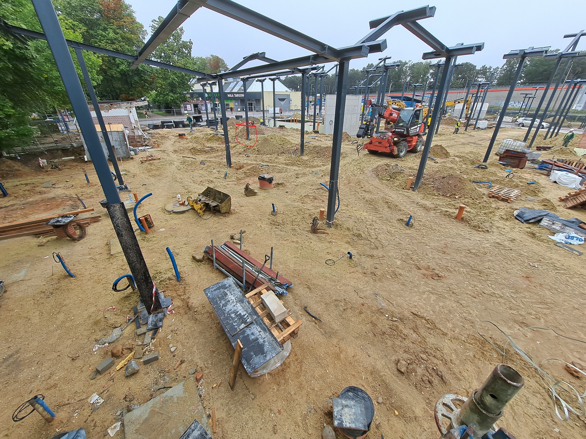 Plac budowy z konstrukcją stalową w trakcie montażu, widoczny sprzęt budowlany i materiały. Ujęcie z góry ukazuje postęp prac i przygotowanie terenu pod budowę.