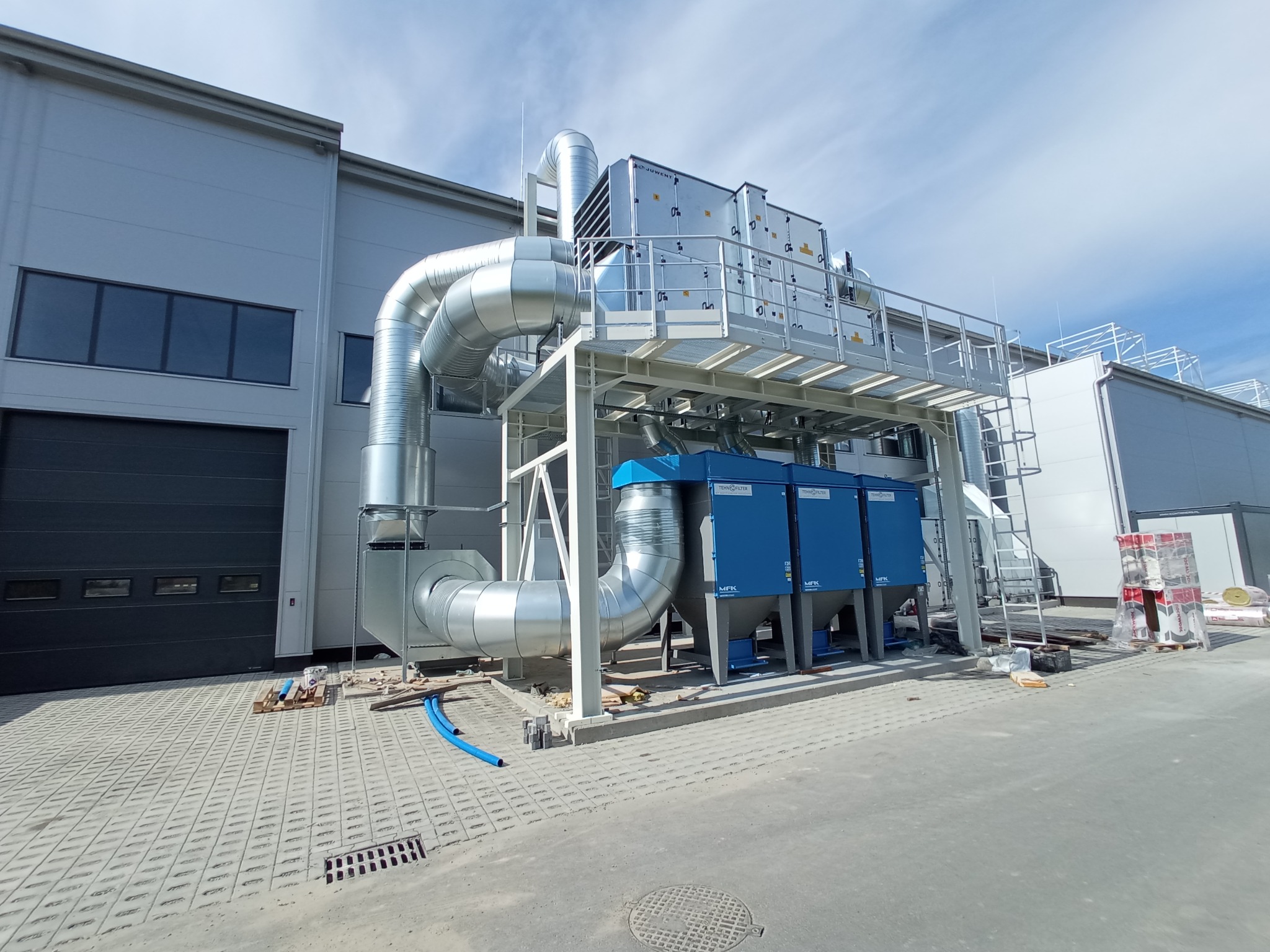 Zewnętrzna instalacja przemysłowa z srebrnymi rurami i niebieskimi filtrami, zamontowana przy szarej hali z bramą garażową, widoczna w słoneczny dzień.