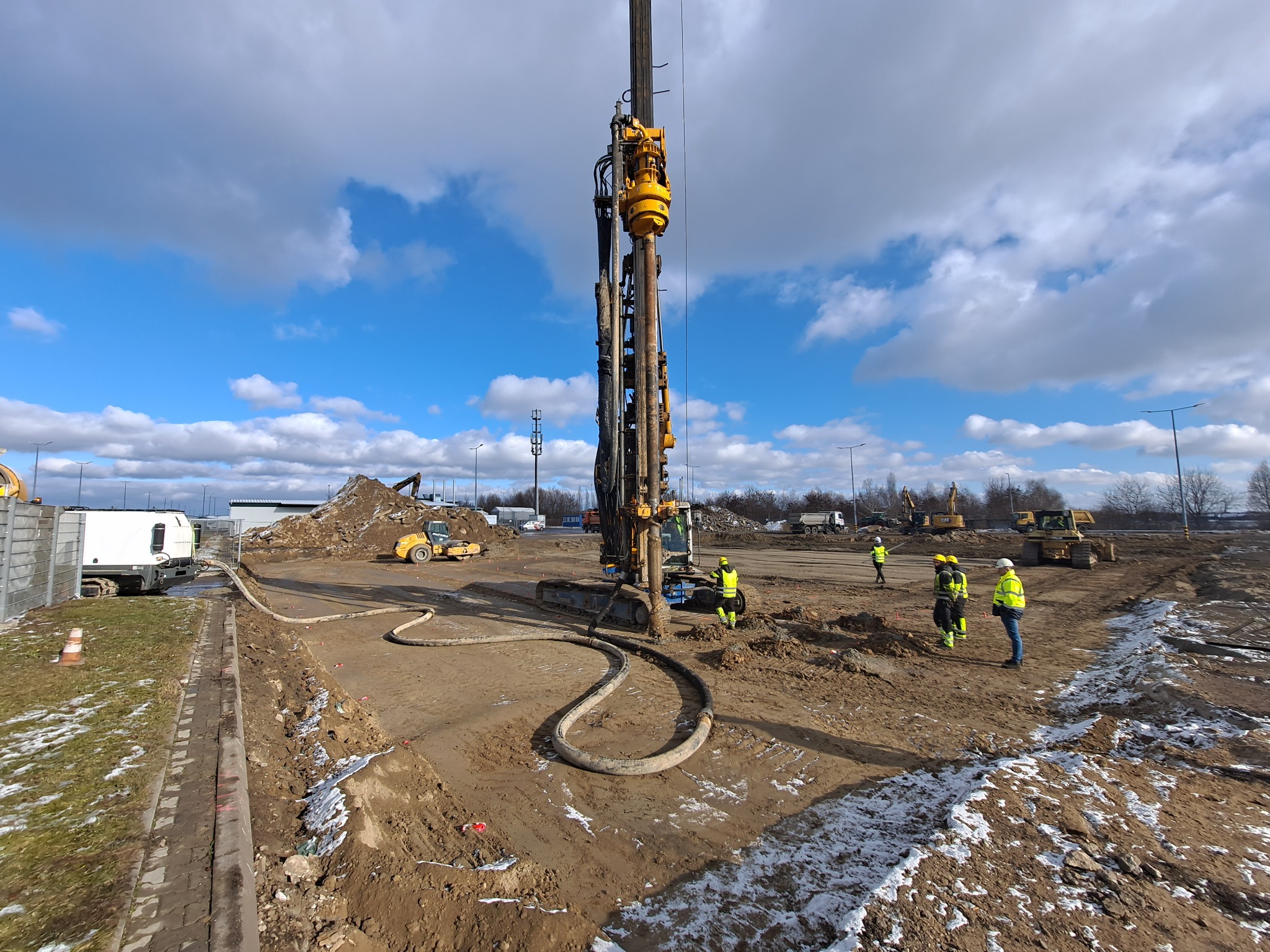 Prace ziemne w toku: ekipa w żółtych kamizelkach nadzoruje wbijanie pali w grunt za pomocą palownicy. W tle widoczny sprzęt budowlany i błękitne niebo z chmurami.