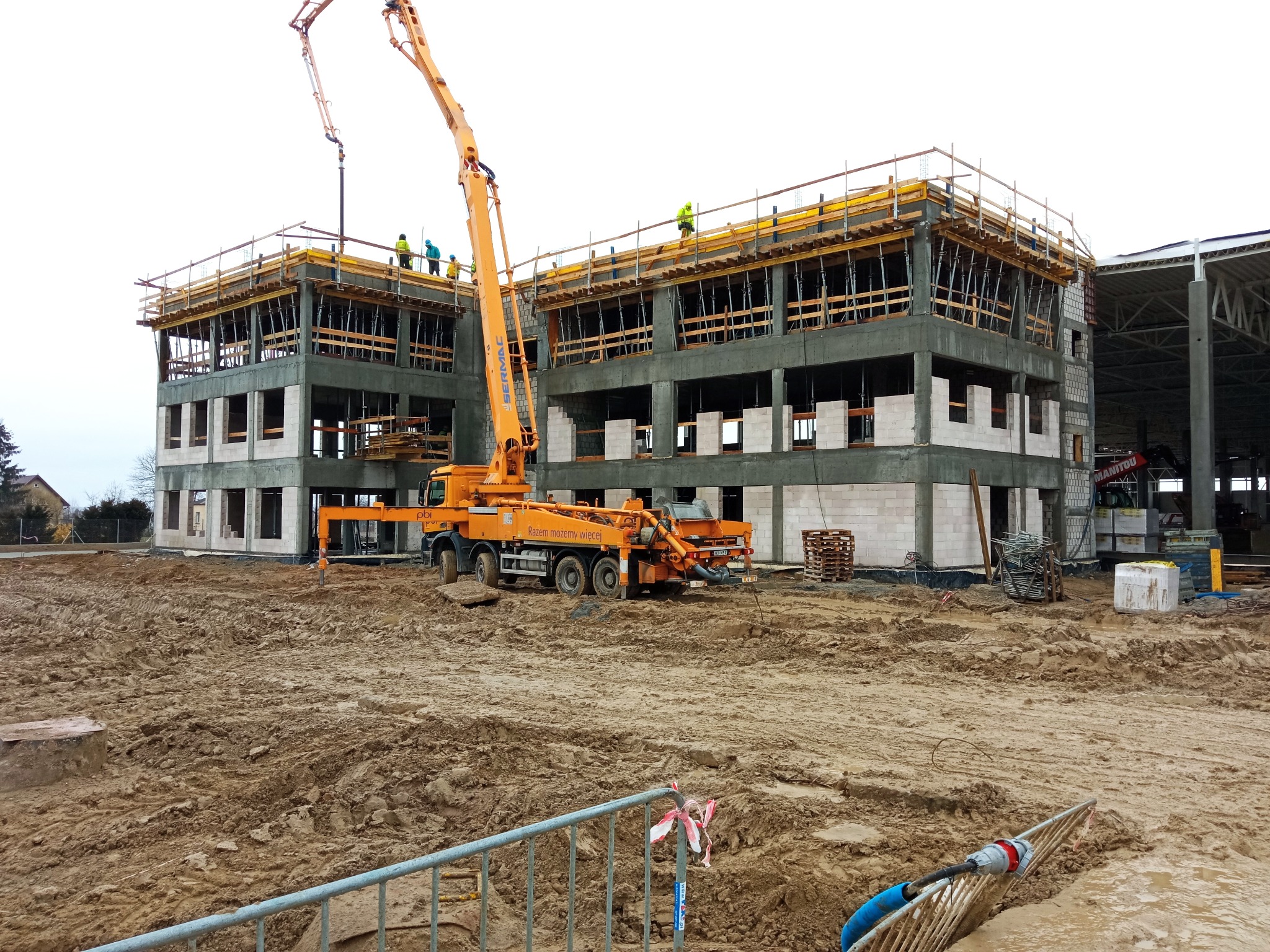 Budowa dwukondygnacyjnego budynku z betonu komórkowego. Na górze szalunki i pracownicy w kamizelkach. Pomarańczowa pompa do betonu w akcji. Błotnisty teren budowy.