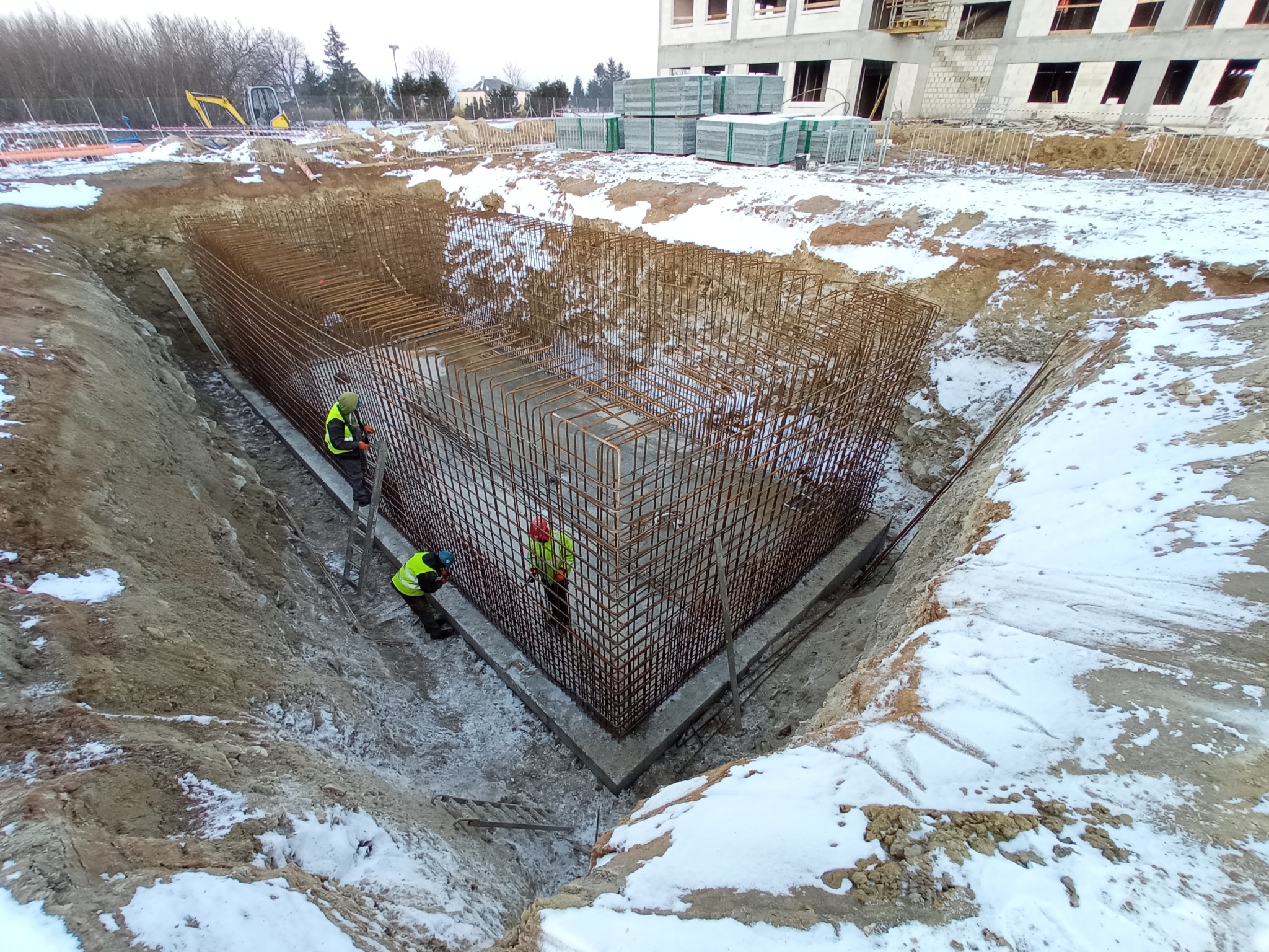 Zbrojenie fundamentu w wykopie, widoczne z góry. Dwóch pracowników w kamizelkach odblaskowych przy zbrojeniu. W tle budynek w stanie surowym i zaspy śniegu.