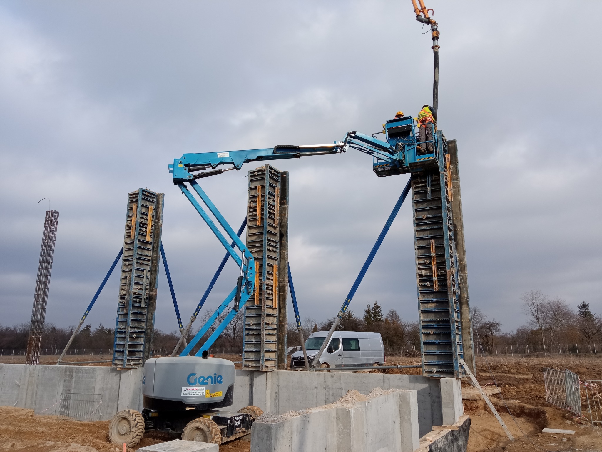 Wznoszenie żelbetowych słupów na fundamencie z użyciem szalunków i podnośnika Genie. Dwóch pracowników w kaskach nadzoruje proces. W tle widoczny bus i zbrojenie.