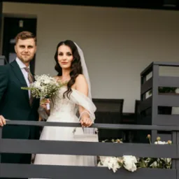 Para młoda: panna młoda w sukni z bukietem, pan młody w zielonym garniturze. Ujęcie z balkonu z kwiatami. Naturalne światło, ciepła tonacja.