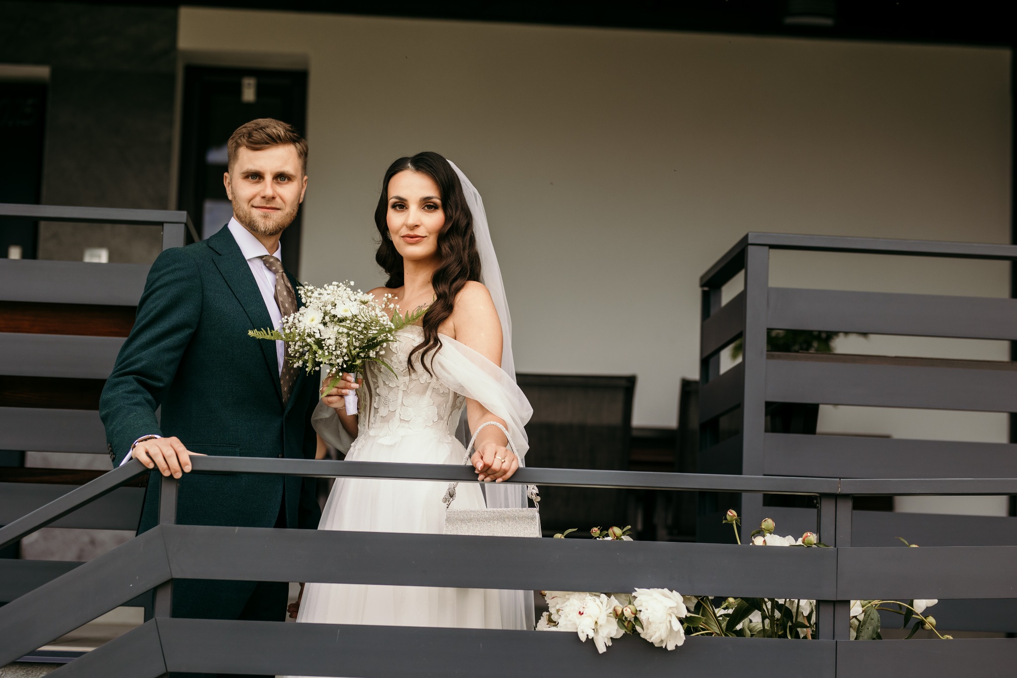 Para młoda: panna młoda w sukni z bukietem, pan młody w zielonym garniturze. Ujęcie z balkonu z kwiatami. Naturalne światło, ciepła tonacja.