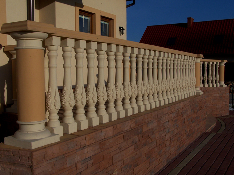 Balustrada balkonowa z prefabrykowanych kolumn betonowych z dekoracyjnym motywem liści, zamontowana na ścianie z cegły klinkierowej, widok z zewnątrz.