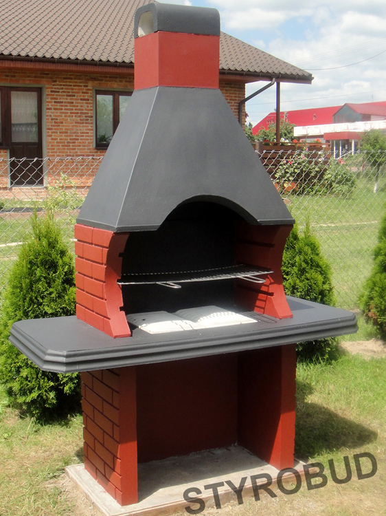 Murowany grill ogrodowy z czerwonej cegły i szarą konstrukcją kominową, z blatem roboczym, na tle zielonego trawnika i domu z czerwonej cegły.