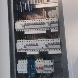 Rozdzielnia elektryczna z zainstalowanymi bezpiecznikami, widoczne przewody i listwy zaciskowe. Szara obudowa w białej ścianie.