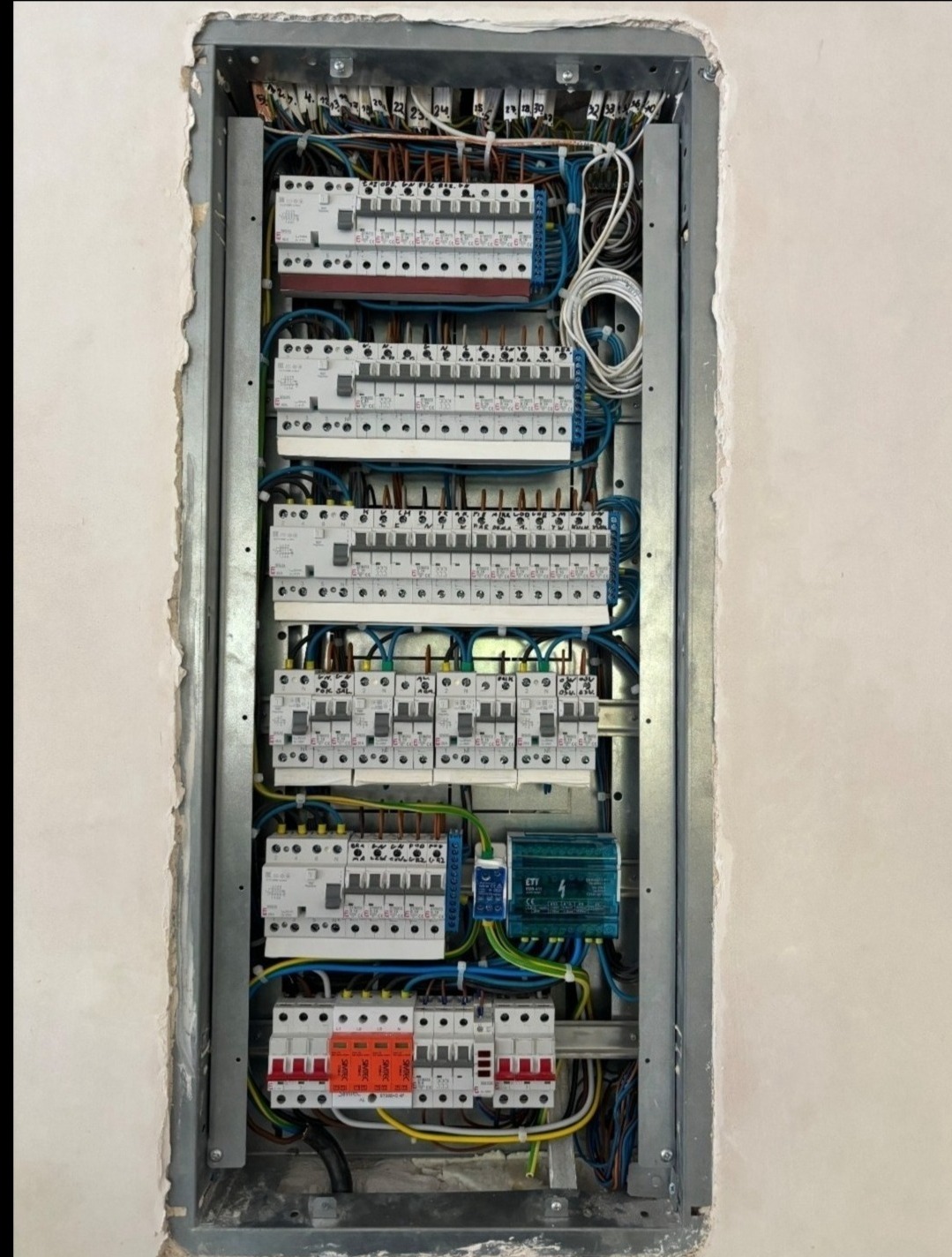 Rozdzielnica elektryczna w metalowej obudowie, zamontowana w ścianie. Widoczne liczne bezpieczniki, przewody i zabezpieczenia. Ściana wokół surowa, przygotowana do wykończenia.