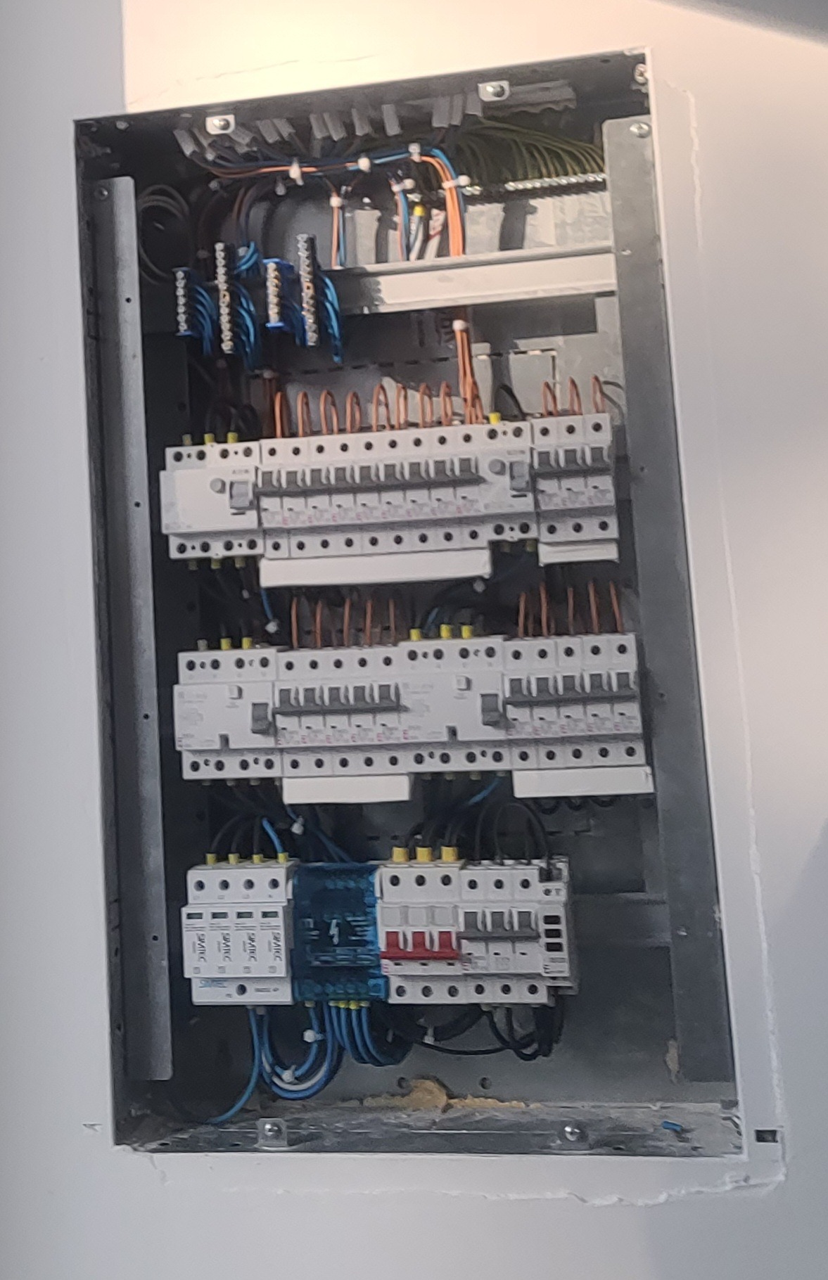 Rozdzielnia elektryczna z zainstalowanymi bezpiecznikami, widoczne przewody i listwy zaciskowe. Szara obudowa w białej ścianie.