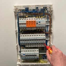 EL-Bro - Otwarta skrzynka elektryczna z bezpiecznikami, widoczny fragment dłoni z wkrętakiem podczas prac instalacyjnych. Kable uporządkowane wewnątrz skrzynki.