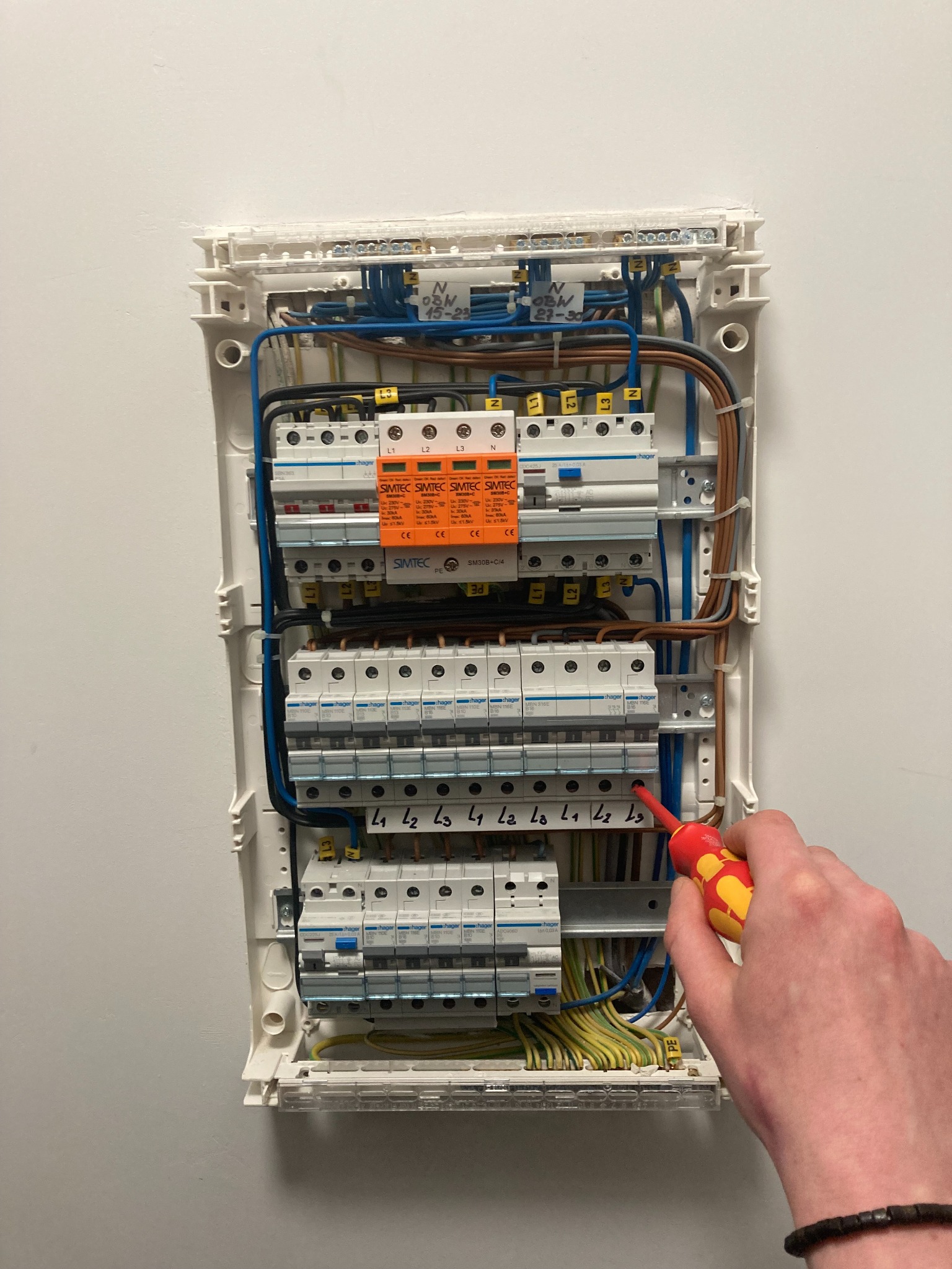 Otwarta skrzynka elektryczna z bezpiecznikami, widoczny fragment dłoni z wkrętakiem podczas prac instalacyjnych. Kable uporządkowane wewnątrz skrzynki.