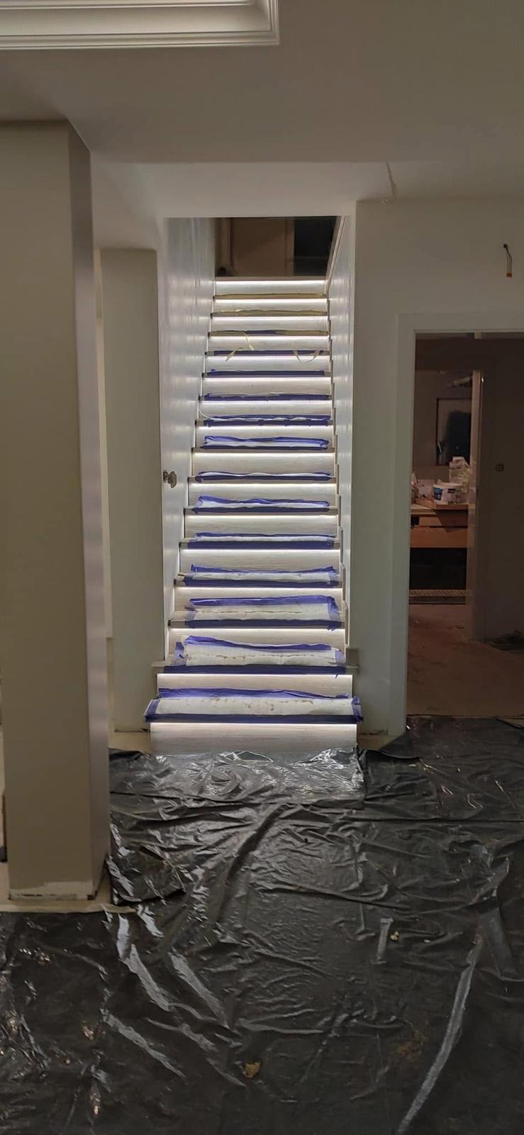 Białe schody z podświetleniem LED, zabezpieczone folią malarską. Widok z dołu, w tle widoczne pomieszczenie w trakcie remontu. Nowoczesne oświetlenie schodowe.