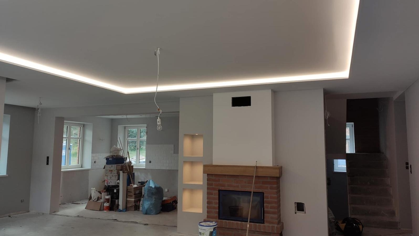 Salon w trakcie remontu z podwieszanym sufitem i oświetleniem LED. Widoczny kominek z cegły i drewnianym blatem. Częściowo widoczne schody i okna.