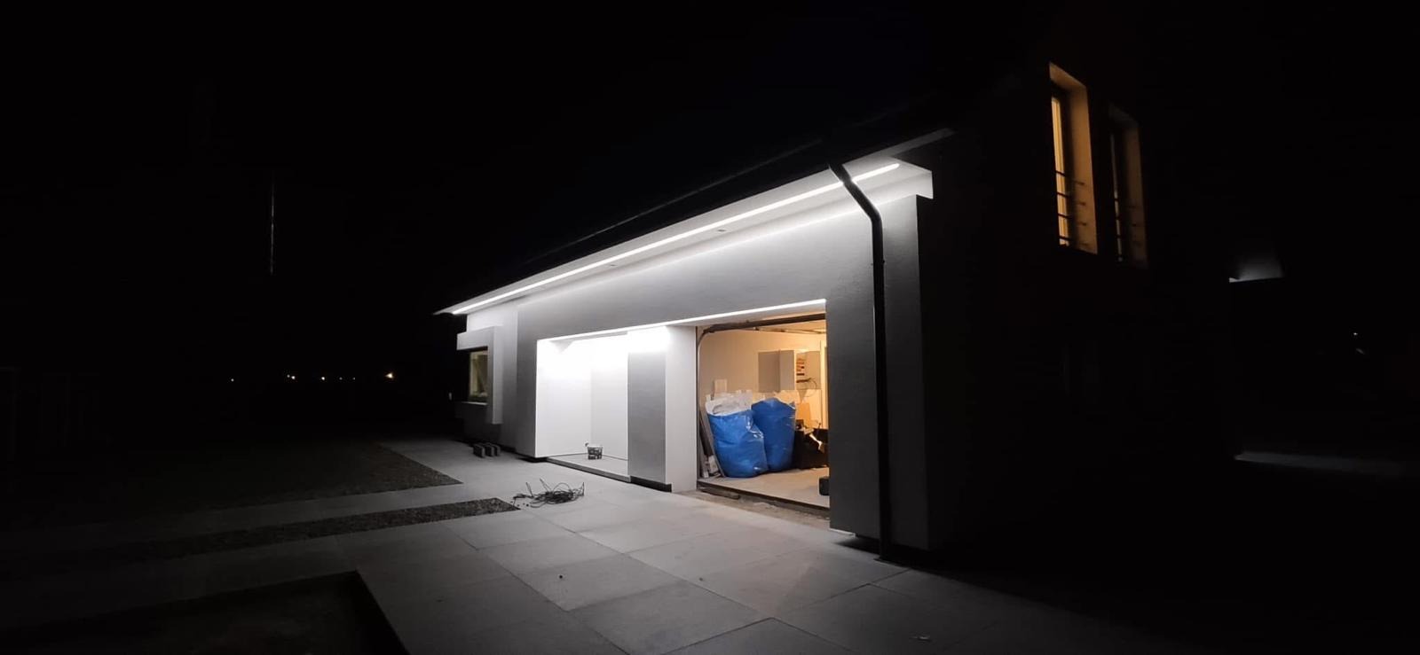 Nowoczesny dom z garażem oświetlonym listwami LED, widoczne dwa worki w garażu, nocne ujęcie z zewnątrz.