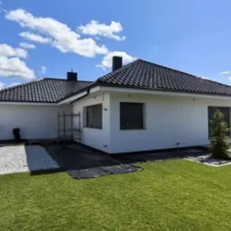 Biała elewacja domu jednorodzinnego z ciemną dachówką, widok z boku, ogród z trawnikiem i ścieżką z kamienia. W tle błękitne niebo z chmurami.