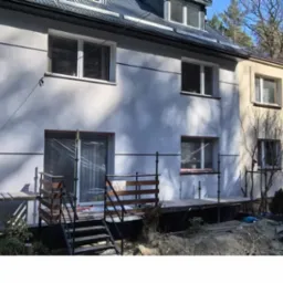 Dwukondygnacyjny dom w trakcie tynkowania elewacji, z widocznymi rusztowaniami i świeżo otynkowaną fasadą w odcieniach bieli i beżu, w słoneczny dzień.